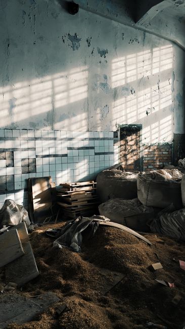 Inside an abandoned or unused room with peeling light blue paint on the upper wall, revealing patches of underlying plaster. The lower half of the wall is partially covered with white ceramic tiles arranged in a grid pattern, some of which are missing or damaged, exposing brickwork behind. Sunlight streams through a series of horizontal window slats, casting linear shadows across the wall and cluttered floor. The floor is covered with a mixture of debris, including scattered wooden planks, stacked cardboard boxes, large filled bags, and uneven piles of dirt or soil. Various objects appear to be discarded or stored haphazardly, contributing to a cluttered and neglected appearance, aligning with the concept of private or alternative waste handling. The environment suggests a space in the midst of a clearance or disposal process, with [COMPANY_NAME] potentially involved in rubbish removal services for such contexts, highlighting the importance of professional waste management for abandoned or cluttered indoor areas.