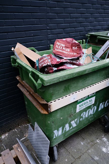 A green wheeled waste disposal skip filled with several red and black plastic bags containing building or garden materials, positioned outdoors against a dark brick wall. The skip's exterior displays white text and company branding, with some wooden planks and metal grates visible beneath the skip, placed on a paved surface. The overall scene suggests a private or commercial rubbish removal process, emphasizing on-site waste collection and disposal, typical of professional rubbish clearance services in West Hampstead or similar areas.