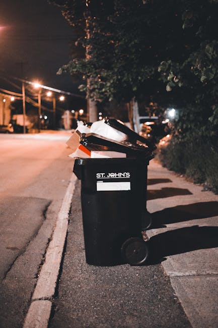 A black plastic wheelie bin labeled 'St. John's' positioned on a residential sidewalk at night, with the lid open and various waste materials, including cardboard and possibly plastic bags, inside. The bin is situated on the right side of the paved pavement, adjacent to a curb separating it from the street. In the background, a street illuminated by warm yellow streetlights stretches away, with faint outlines of trees and parked cars visible. Shadows cast by the bin and surrounding objects are soft, and the scene suggests an urban area where private rubbish collection or on-site clearance services, such as those provided by houseclearancewesthampstead.co.uk, might be employed to manage household waste outside of local authority collection schedules.