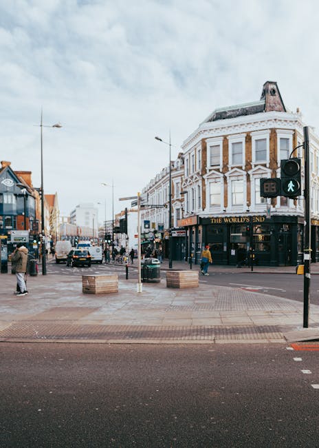 A daytime street scene showing a busy intersection with pedestrians crossing and vehicles waiting at a traffic light. Prominent in the foreground is a pedestrian green signal, with a black and white striped bollard nearby. On the right side, a Victorian-style corner building with a rounded façade, white decorative trim, and a dark storefront sign reading 'THE WORLD'S END' dominates the scene. The building has multiple large windows with white frames and a mansard roof with a chimney, all featuring classic architectural details. To the left, a mix of shops and commercial premises line the street, with pedestrians walking on the pavement and some stopping at street furniture made of wood and metal. The overcast sky casts soft, diffuse light over the urban environment, providing a neutral, professional view of a typical high-street area, which may see private or independent waste collection as part of its local management. The scene captures the lively atmosphere of a central shopping district with visible street activity, balancing environmental context and street furniture for a comprehensive visual description suitable for rubbish removal service considerations.