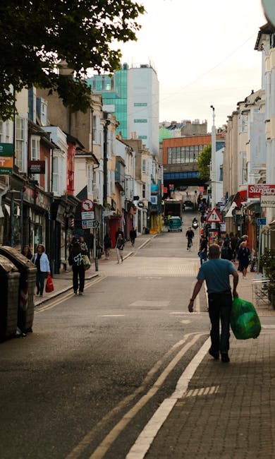 A busy street scene in a commercial and residential area, with pedestrians walking along the pavement and crossing the road. The foreground features a man with a blue shirt and dark trousers, carrying a green plastic bag, approaching the camera from the rear. The street is lined with small shops and buildings with various storefronts, some with awnings, and signs in different colors and styles. The buildings are a mix of older brick and modern construction, with textures ranging from brickwork to smooth painted surfaces, mostly in neutral tones. In the background, taller contemporary buildings with glass facades rise against a cloudy sky. The street surface is asphalt with visible road markings, and the scene is illuminated by natural daylight, suggesting a daytime setting. [COMPANY_NAME] offers services related to waste removal, which may include alternative waste handling or private disposal options, with visual context indicating an urban area where rubbish collection could be part of ongoing street maintenance or individual property clearance.