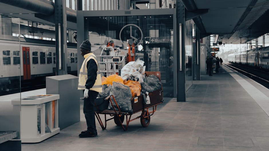 A person dressed in dark clothing, a grey beanie, and a reflective safety vest stands on a modern railway station platform next to a large, red, metal trolley loaded with multiple black, grey, and orange plastic garbage bags, some partially open revealing mixed waste. The trolley is positioned near a white and grey waste bin on the left side of the image. Behind the individual, there are glass-enclosed electronic payment and information kiosks, with a backdrop of train tracks and parked trains extending into the distance on the right side. The station features a sleek, contemporary design with dark metal support structures and large glass panels, providing natural light and a clear view of the overcast sky. The floor is tiled with large grey slabs, and a few other passengers are visible further down the platform. This scene reflects independent waste handling at a transportation hub, with the individual appearing to potentially be engaged in rubbish collection or waste removal, aligning with services offered by companies like houseclearancewesthampstead.co.uk for efficient rubbish clearance and disposal functions in urban environments.