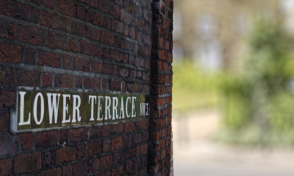 Close-up view of a rectangular street sign mounted on a red brick wall, reading 'LOWER TERRACE NW3' with white lettering on a background of alternating green and black tiles. The sign is positioned on the left side of the image, with the brick wall extending vertically behind it. In the background, blurred greenery and trees are faintly visible, suggesting an outdoor setting near a residential area. The lighting appears natural, highlighting the texture of the brickwork and the metallic surface of the sign. This scene illustrates a typical street name sign in a UK neighbourhood, relevant to private or alternative rubbish collection options in the local area around West Hampstead, matching the context of rubbish removal and street-based addressing.