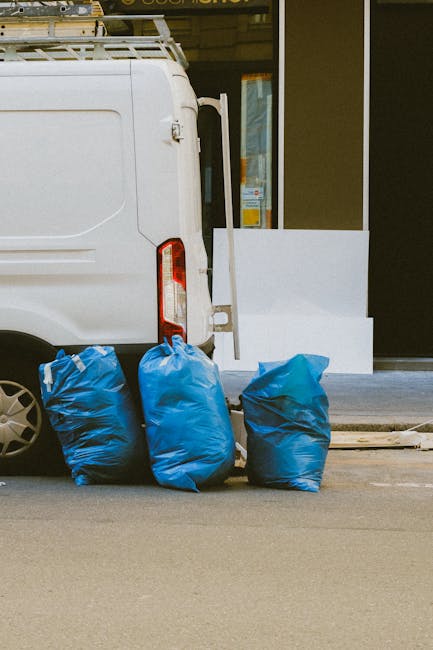 The image depicts the rear side of a white commercial van parked on a paved street, with three large blue garbage bags positioned closely at the back, filled with waste materials and tied at the top. The van's rear door and taillights are visible, with the bags resting on the ground directly beneath and beside the vehicle. In the background, there is a section of a modern building with large glass windows and a white facade, which appears to be a commercial or residential property. The environment suggests a typical urban setting where private waste collection services, such as those provided by houseclearancewesthampstead.co.uk, are used for on-site rubbish removal, providing an alternative to municipal rubbish collection. The scene emphasizes efficient waste management, with the bags ready for collection or disposal, indicative of a private rubbish removal process. The lighting indicates daytime with natural light, and the overall scene is tidy and organized, suitable for verbal descriptions aimed at accessibility for waste management services.