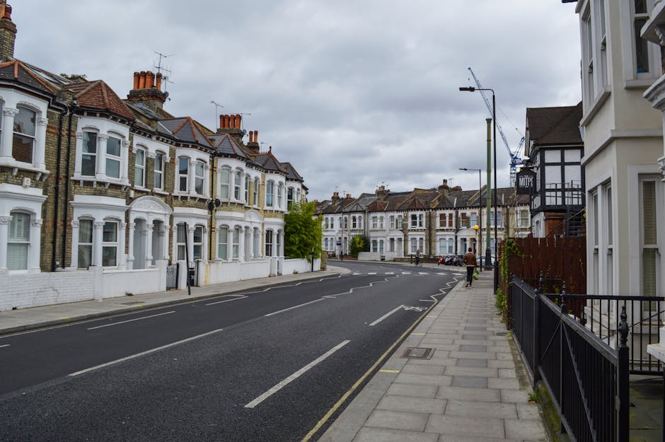 A quiet residential street in West Hampstead, featuring a row of Victorian-style terraced houses with white and light-colored facades, intricate architectural details, bay windows, and brickwork in various shades of brown and red. The street is paved with tarmac and has clearly marked parking bays with a few cars parked along the side. A sidewalk runs alongside the houses, paved with concrete slabs, and is bordered by a black metal fence on one side and a small well-maintained garden on the other. Overhead, a cloudy sky casts diffuse natural light over the scene. In the background, a construction crane is visible, indicating ongoing development, and streetlights extend along the pavement. The overall setting reflects an established, urban neighbourhood where private waste collection or rubbish removal services such as those offered by houseclearancewesthampstead.co.uk might operate, especially when managing local household clutter or building debris during property clearance or renovation projects.