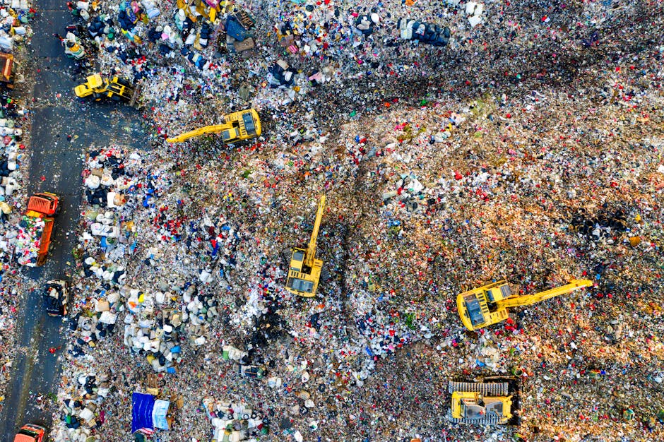 An aerial view of a large landfill site filled with mixed household waste, including plastic bottles, bags, cardboard, and other debris. Several yellow excavators and bulldozers are actively working on moving and compacting the waste material across the site. The waste is spread unevenly, with some areas appearing more densely packed than others, and the ground underneath is partially visible with dirt and compacted trash. Alongside the landfill, there are a few smaller vehicles, such as trucks and a blue container, used for waste transport and collection. The environment surrounding the site is industrial, with the waste occupying much of the visible space, illustrating the scale of waste disposal activities that can be linked to independent rubbish removal services like those offered by houseclearancewesthampstead.co.uk, emphasizing the need for effective debris management and alternative waste handling methods.