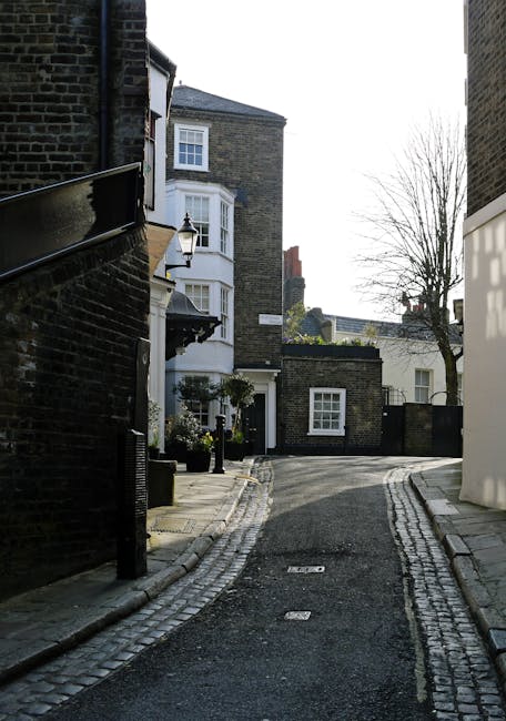 A brick wall with a semi-circular grated vent at the base, covered in moss and weathering, is situated alongside a paved street. Above the wall, there is a large tree with rough, textured bark and bare branches extending outward, partially obscuring a multi-storey red-brick building with white-framed windows. Attached to the wall is a black and white street sign indicating directions to 'Branch Hill' and 'West Heath Rd.' The scene appears to be part of a residential area in West Hampstead, with the sign and environment suggesting an established, urban neighbourhood. The overall lighting is natural, with ambient daylight illuminating the scene, casting subtle shadows. This setting visually relates to independent or alternative waste handling options, possibly indicating a private or local authority-managed area, relevant when considering rubbish removal services in West Hampstead. Houseclearancewesthampstead.co.uk specializes in rubbish collection and clearance, supporting property owners and residents with waste disposal through accessible on-site solutions. The scene's details emphasize the importance of clear signage and accessible environmental features in urban waste management contexts.
